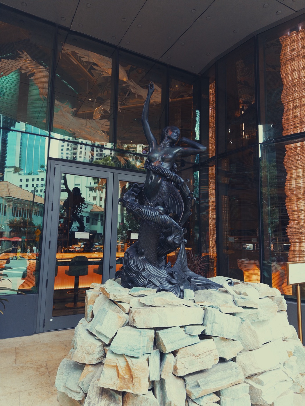 A dark bronze sculpture on stacked stone in front of a glass lobby